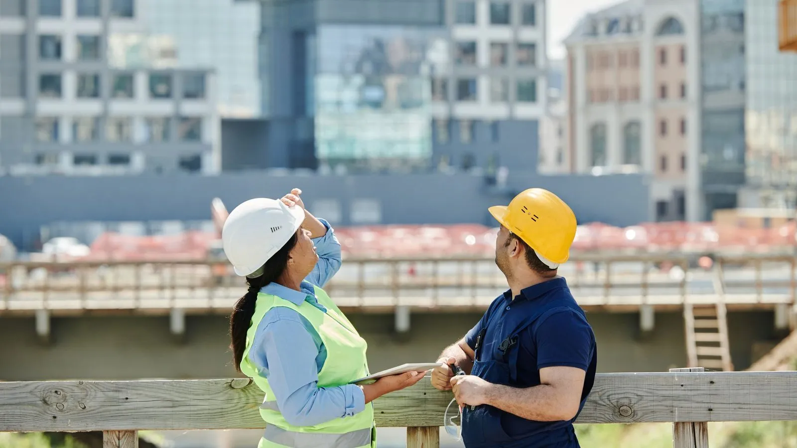 Two people in hard hats at a fence, looking up at a building